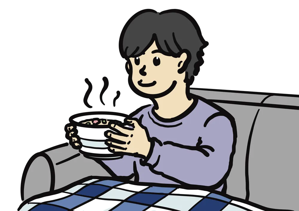 　ソファに座って、チェックのブランケットを膝に乗せた男性が湯気の出たスープを持っている様子